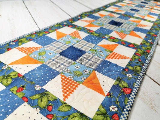 This long table runner features a strawberry focal fabric. A variety of pretty blue fabrics are complimented by orange and cream creating a lovely, summery table runner.