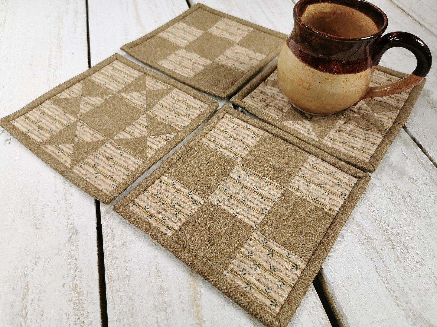 Four beige patchwork coasters are grouped close together.  A coffee mug sitting on one shows scale. Two coasters are pieced in a star pattern and the other two are nine patches. 