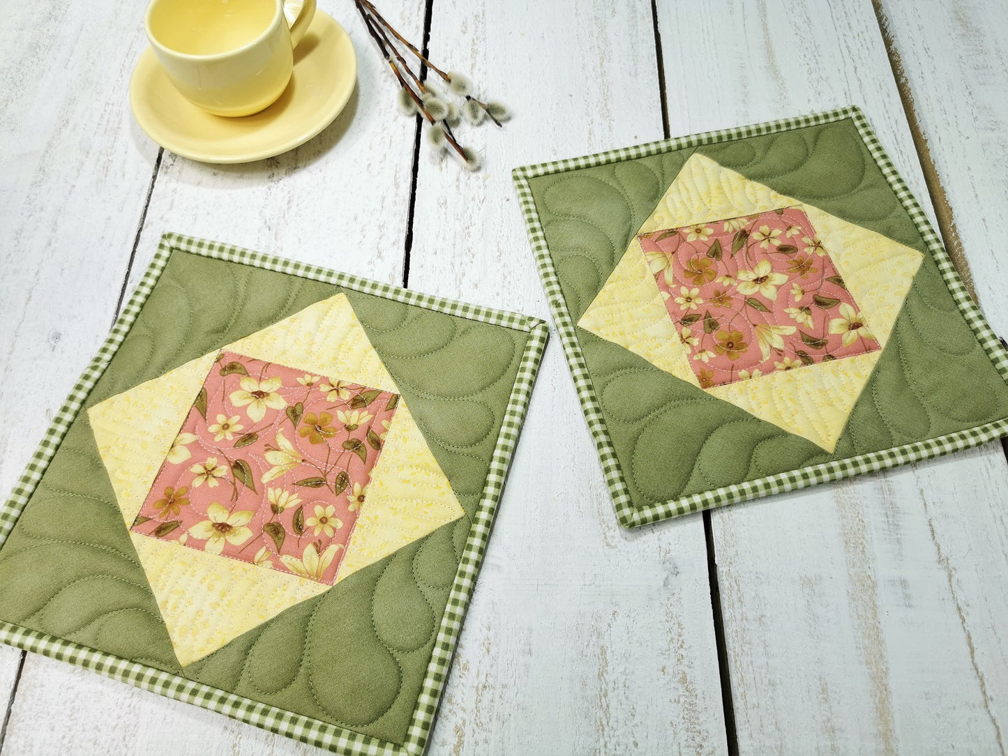 two pretty green and yellow patchwork potholders  with a pink floral focal fabric. Custom quilting adds nice texture. Green gingham binding. Background has small yellow teacup with a few pussywillow branches showing scale.