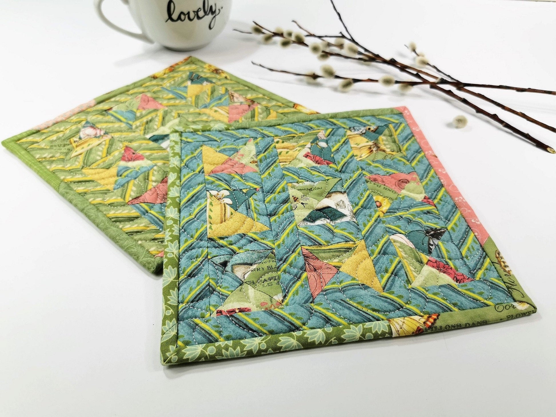 Two scrappy patchwork potholders shown on a white table with a mug and pussy willows in the background. Potholder colors are pastel shades of blue, green, pink, yellow .