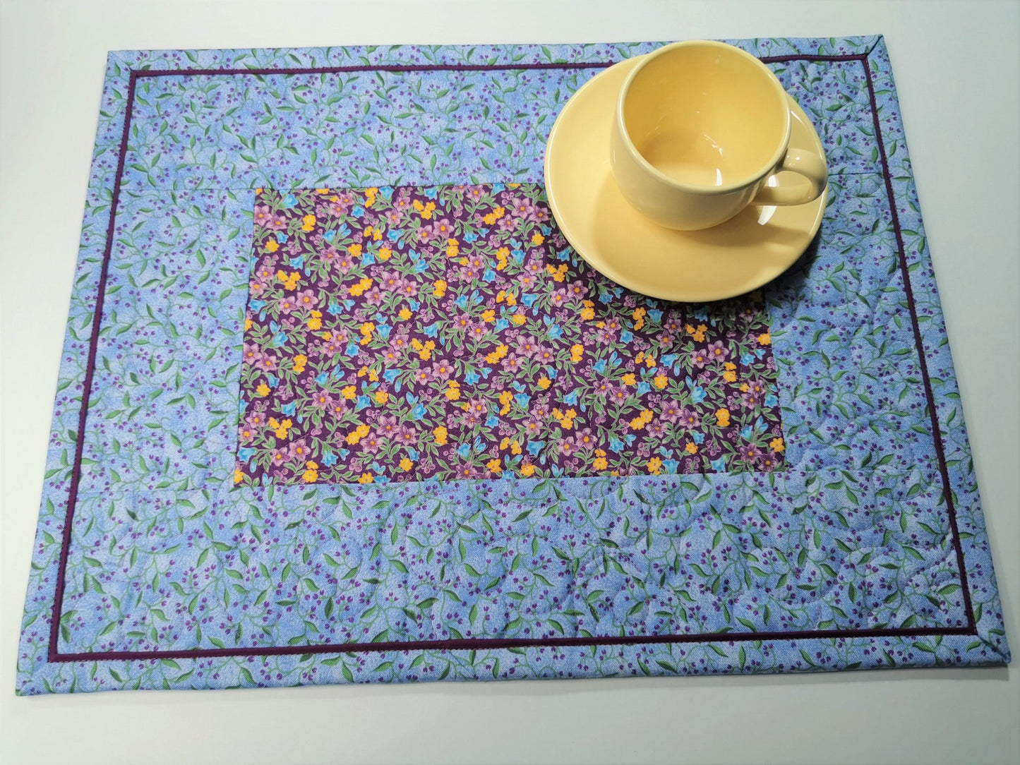 Overhead view of the placemat shown with a small yellow tea cup. The placemats are predominantly a pretty sky blue with purple as a secondary color. The purple floral print in the center has golden yellow flowers scattered throughout. A key feature of the placemats is the flanged binding which gives a pretty framing effect.