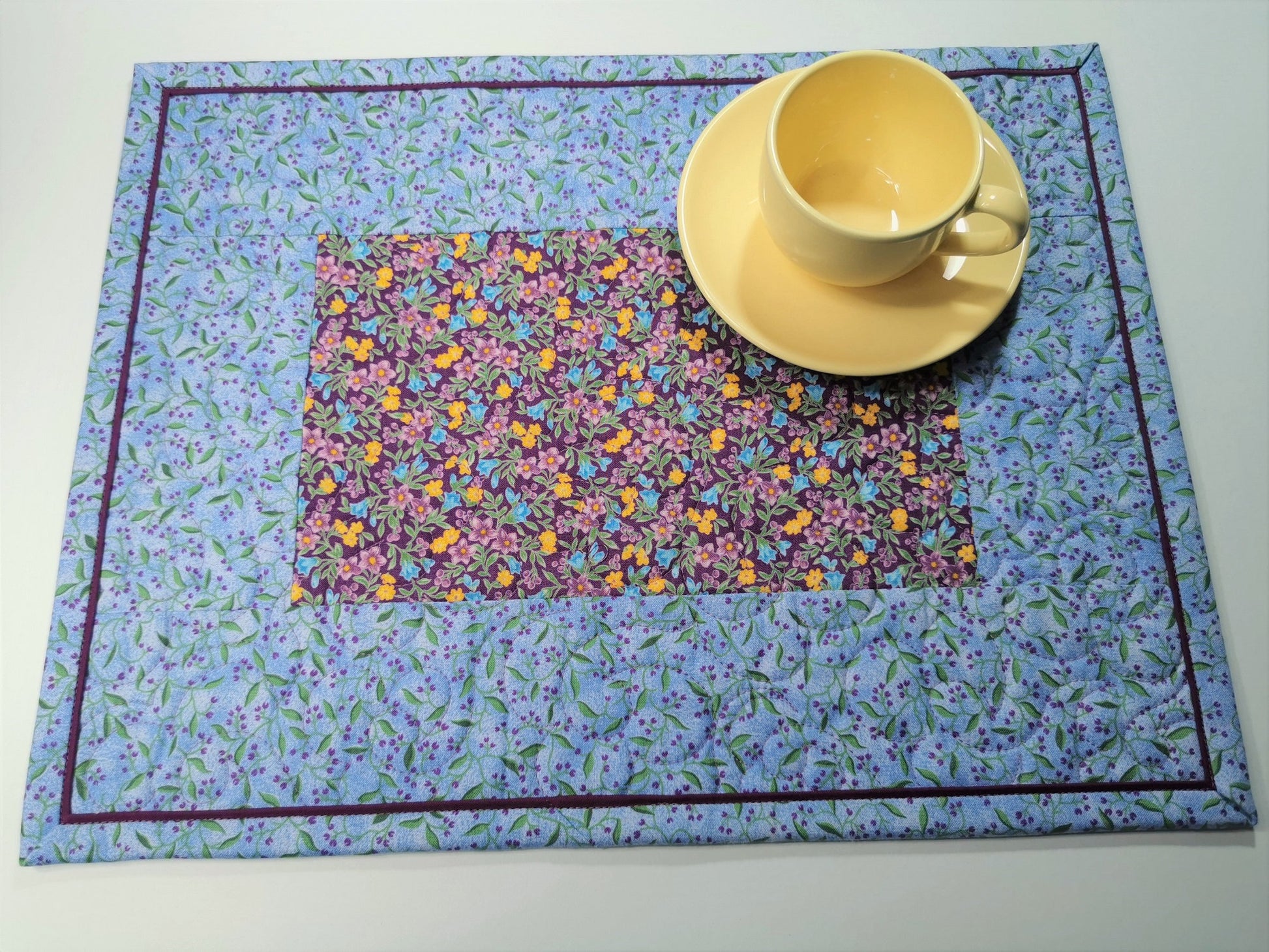 Overhead view of the placemat shown with a small yellow tea cup. The placemats are predominantly a pretty sky blue with purple as a secondary color. The purple floral print in the center has golden yellow flowers scattered throughout. A key feature of the placemats is the flanged binding which gives a pretty framing effect.